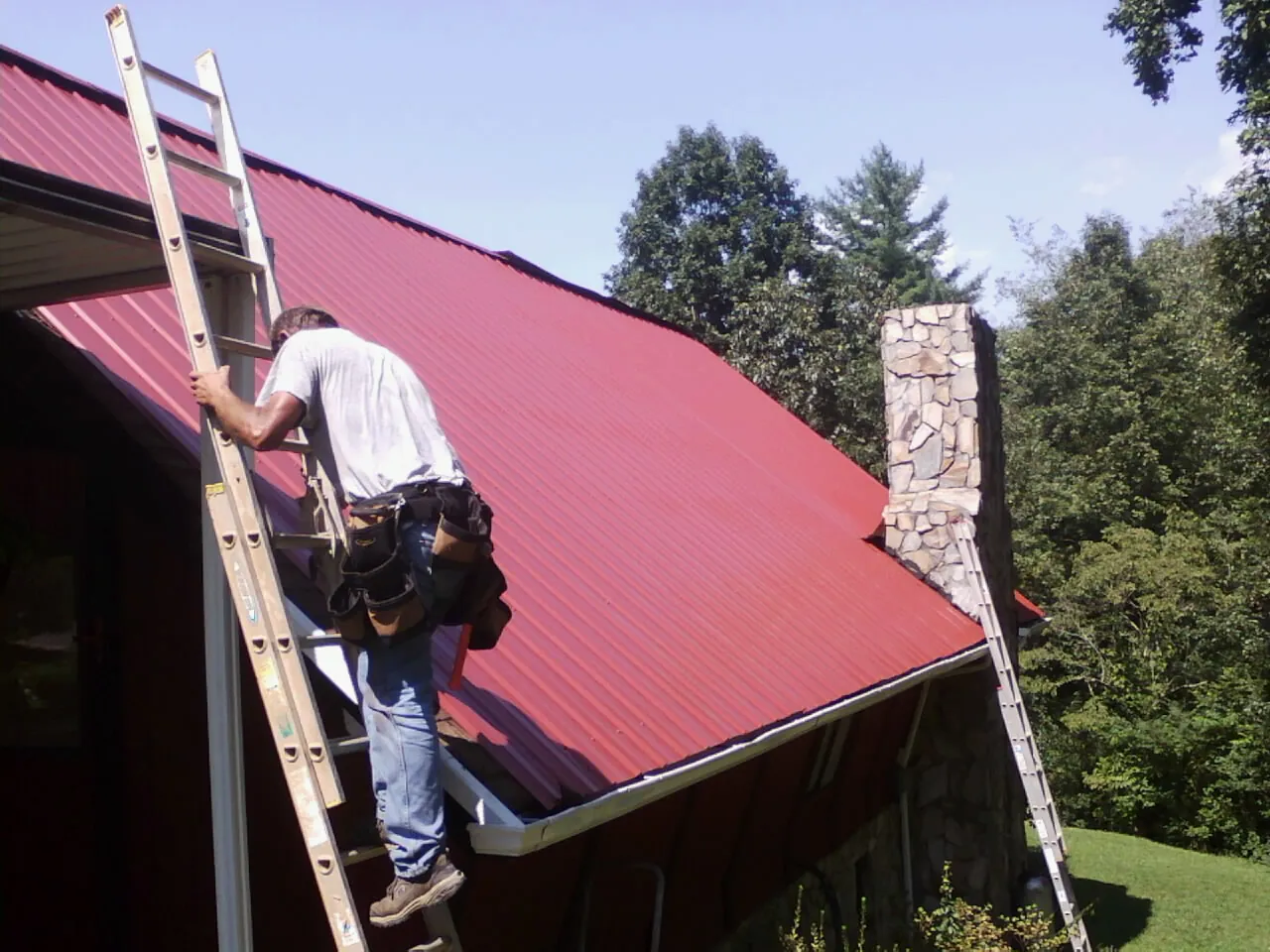 Roofer on a ladder beside a red metal roof and stone chimney — Soffit & Fascia Repair by Roofing Brothers in Brookshire